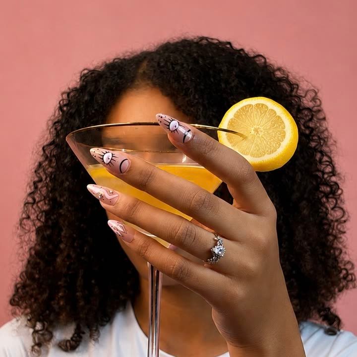 Evil Eye press-on nails, pink shimmer with black line art eyes, held around a martini glass