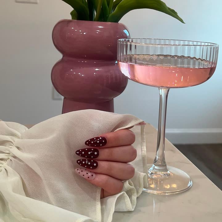 Merlot Dot press-on nails, deep burgundy with white polka dots, beside a glass of rose